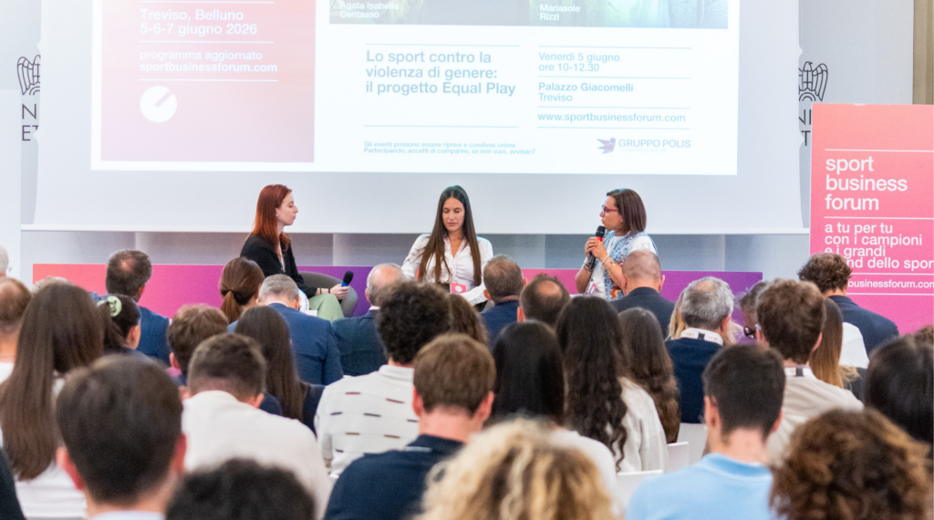 Una platea di spalle assiste a un panel di tre relatrici sul palco durante lo Sport Business Forum 2026. Sullo sfondo, un grande schermo mostra il titolo dell'evento "Lo sport contro la violenza di genere: il progetto Equal Play.